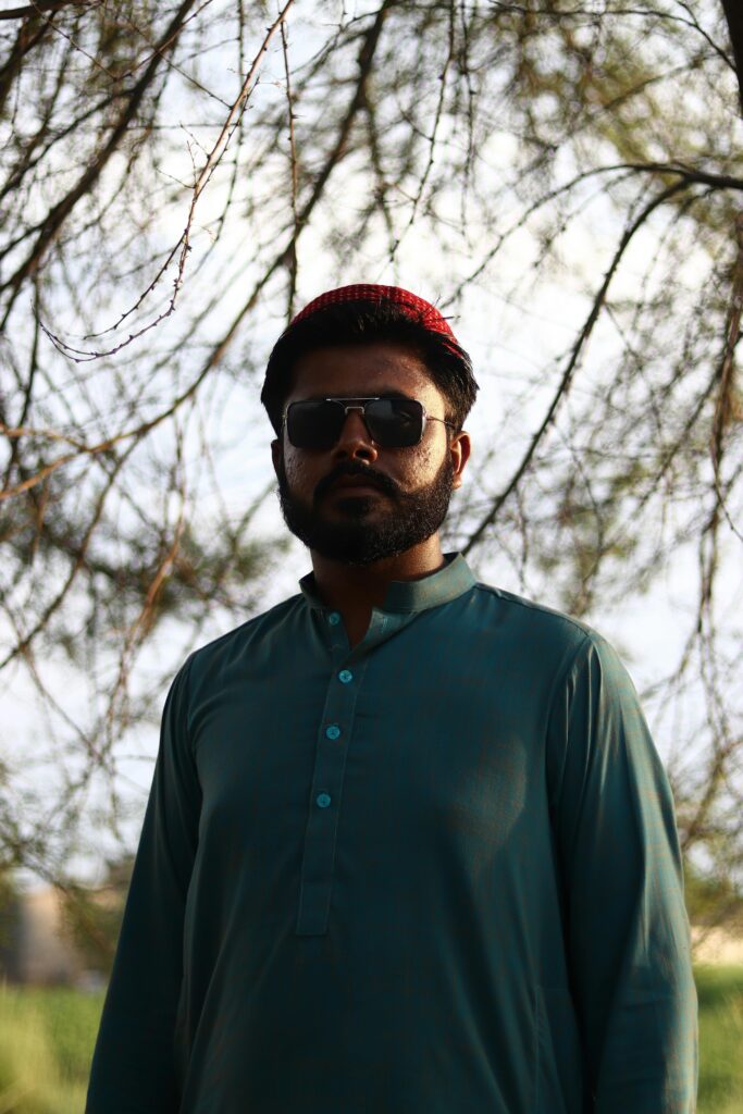 Home A striking portrait of a man wearing traditional clothing and sunglasses, standing outdoors in nature.
