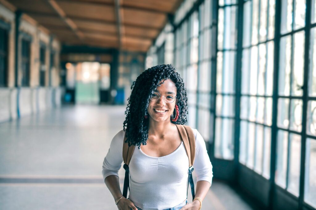 Home A cheerful woman with glasses stands in a bright indoor hallway, carrying a backpack.